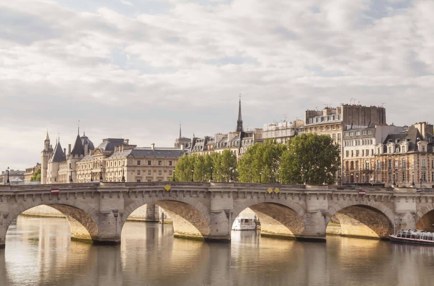 A stone bridge with multiple arches over a calm river, with historic buildings and a tall spire in the background.