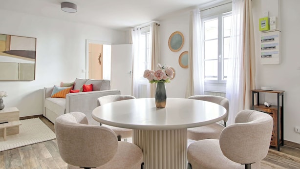 Ein modernes Wohnzimmer mit einem runden Esstisch, beigen Stühlen, einer Couch mit farbenfrohen Kissen und einer Vase mit Blumen.