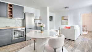 Eine moderne Küche mit integrierten Geräten, ein Essbereich mit rundem Tisch und Stühlen sowie ein Wohnzimmer mit Sofa und Fernseher.