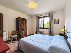 Ein Schlafzimmer mit einem Bett, einem Holzschrank, einem Sessel, einem Fenster mit Vorhängen und einer Heizung.