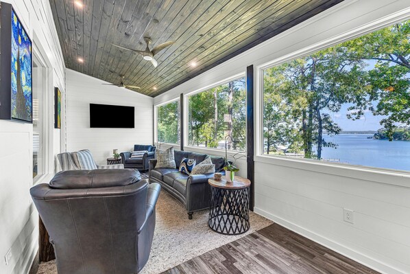 A modern living room with a large window overlooking a lake, a ceiling fan, and a flat-screen TV mounted on the wall. - New! No Worries on Lake Martin 3BR/3BA that sleeps 10, pets OK! (Dadeville)