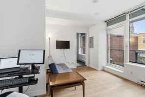 Ein Zimmer mit Schreibtisch, Computer, Stuhl und einem großen Fenster.
