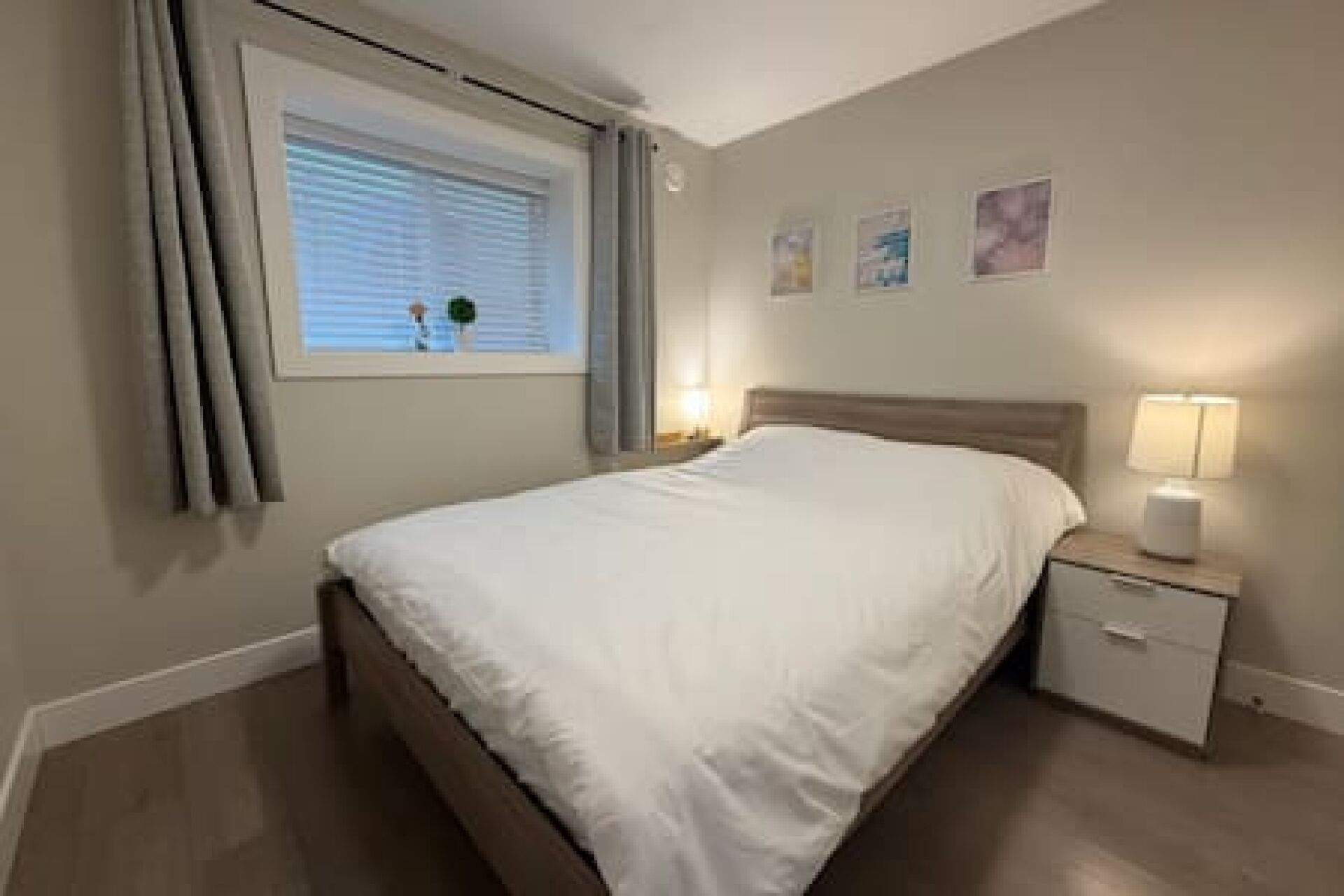 A bedroom with a bed, a nightstand, a window with curtains, and wall art.
