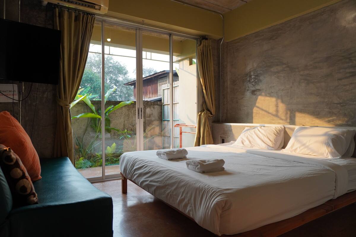 A bedroom with a bed, a sofa, a TV, and a sliding glass door leading to a garden.