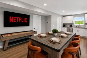 Une cuisine moderne avec des meubles blancs, une table à manger dressée pour six personnes et un grand téléviseur affichant le logo « NETFLIX ».