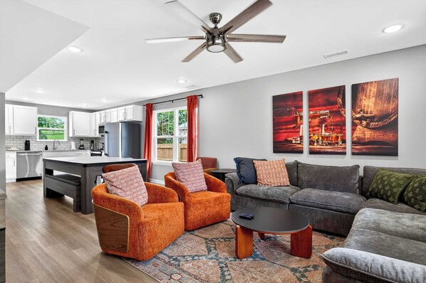 A modern living room with a sofa, armchairs, and a coffee table. There is a kitchen in the background with white cabinets and a refrigerator.