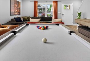 A pool table in a living room with a sofa and cushions, a window with curtains, and a door.