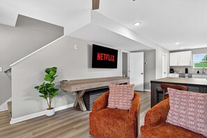 A modern living room with a flat-screen TV, a wooden bench, two orange armchairs, and a potted plant.