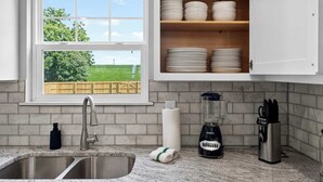 Una cocina con ventana, pileta de acero inoxidable, muebles blancos y una licuadora.
