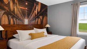 Una cama con ropa de cama blanca y un plaid color mostaza, un cabecero de madera y una pared con un mural con motivos de barriles.