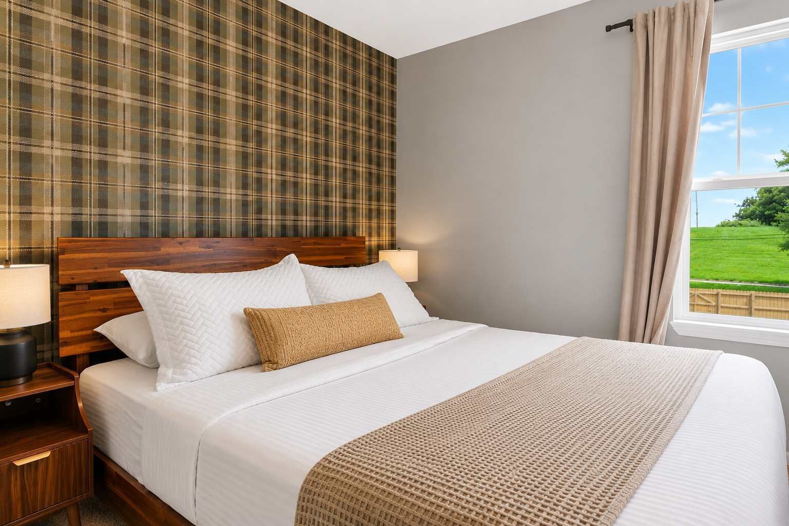 A neatly made bed with a wooden headboard, two bedside lamps, and a view of a green landscape through a window.