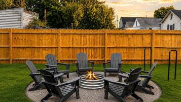 A backyard with a fire pit surrounded by six Adirondack chairs.