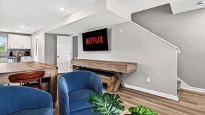 A modern living room with a wooden coffee table, blue chairs, a large TV screen displaying 'NETFLIX,' and a staircase leading to another level.