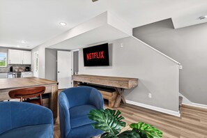 Una sala de estar moderna con una mesa de centro de madera, sillas azules, una gran pantalla de televisión que muestra 'NETFLIX' y una escalera que sube a otro nivel.
