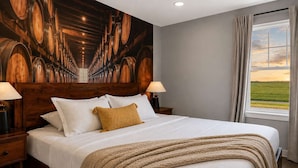 A hotel room with a large bed, wooden headboard, two bedside lamps, a window with a view of the countryside, and a mural of wine barrels on the wall.