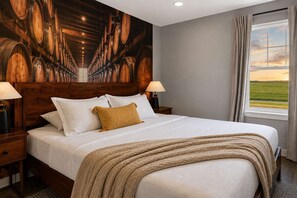 Una habitación de hotel con una cama grande, cabecera de madera, dos lámparas de noche, un ventanal con vista al campo y un mural de barricas de vino en la pared.