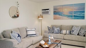 Ett vardagsrum med två soffor, ett soffbord, en takfläkt, en luftkonditioneringsanläggning, en vägglampa och en stor tavla som föreställer en strandvy.