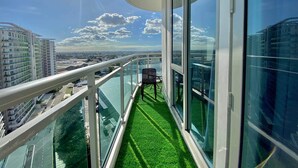 Un balcón con barandilla de vidrio, una silla y vistas a un paisaje urbano y al agua.