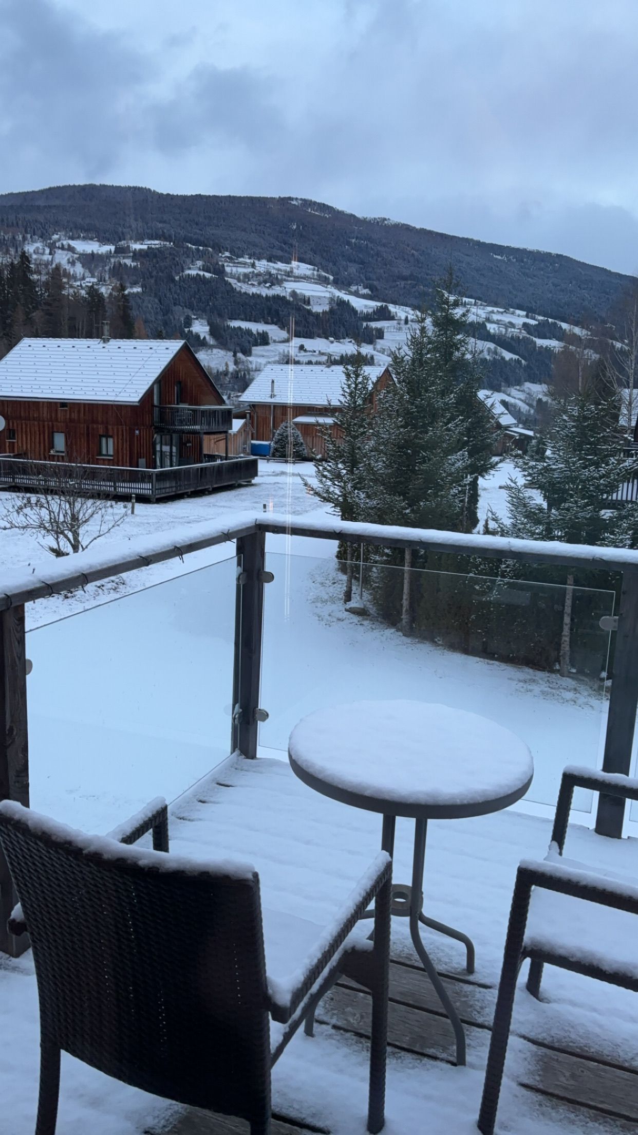A snow-covered deck with a table and chairs, overlooking a snowy landscape with houses and trees.