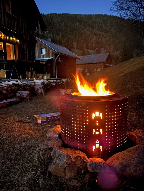 Ein Kaminofen mit Feuer, umgeben von Steinen, in ländlicher Umgebung mit Häusern im Hintergrund.