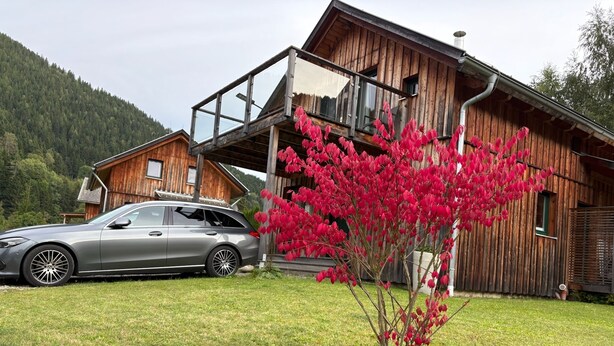 Eine Holzhütte mit einem geparkten Auto davor, ein kleiner Baum mit roten Blättern und eine Rasenfläche.