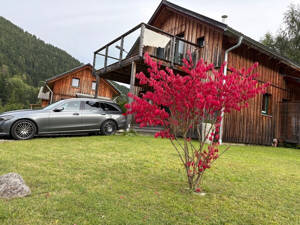 Eine Holzhütte mit einem geparkten Auto davor, ein kleiner Baum mit roten Blättern und eine Rasenfläche.
