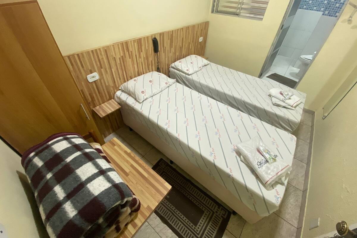 Ein kleines, kompaktes Zimmer mit zwei Einzelbetten, einem Holzschrank, einem kleinen Tisch und einem Fenster mit Jalousien.