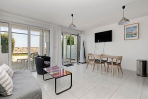 Una sala de estar moderna con sofá, una mesa de centro, una mesa de comedor con sillas, un televisor montado en la pared y una puerta corrediza de vidrio que da a un área exterior.