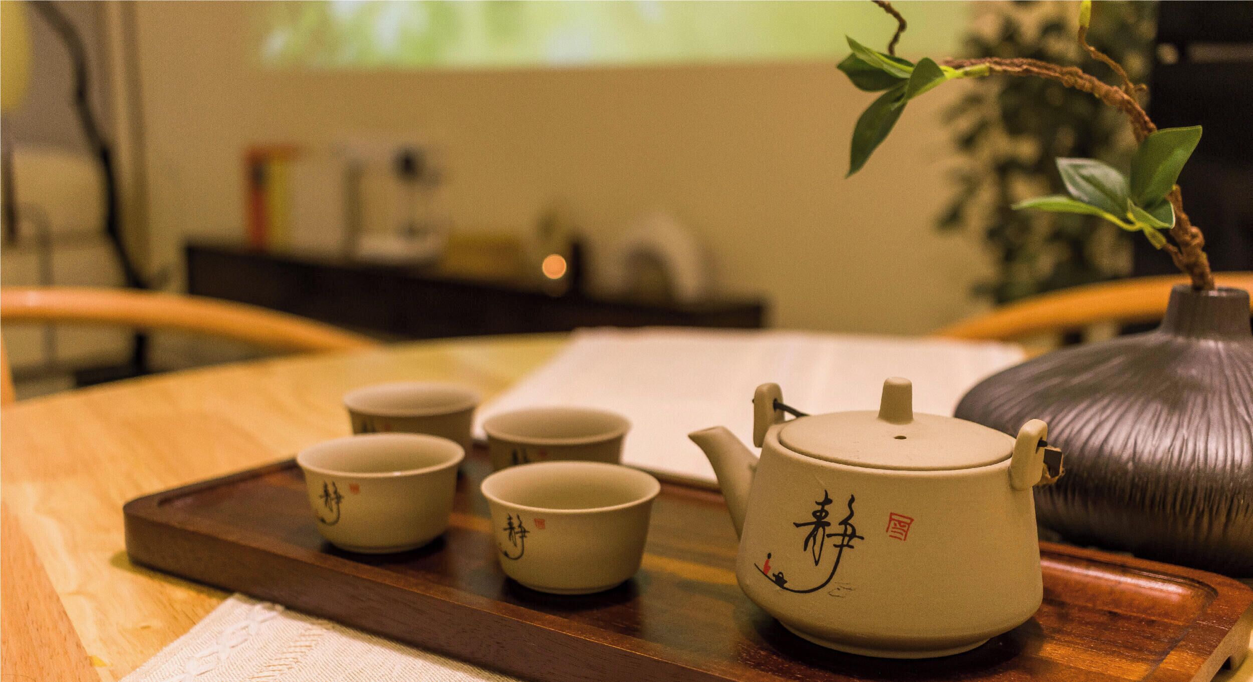 A teapot with four cups on a wooden tray, a vase with a plant, and a blurred background with a projector screen.