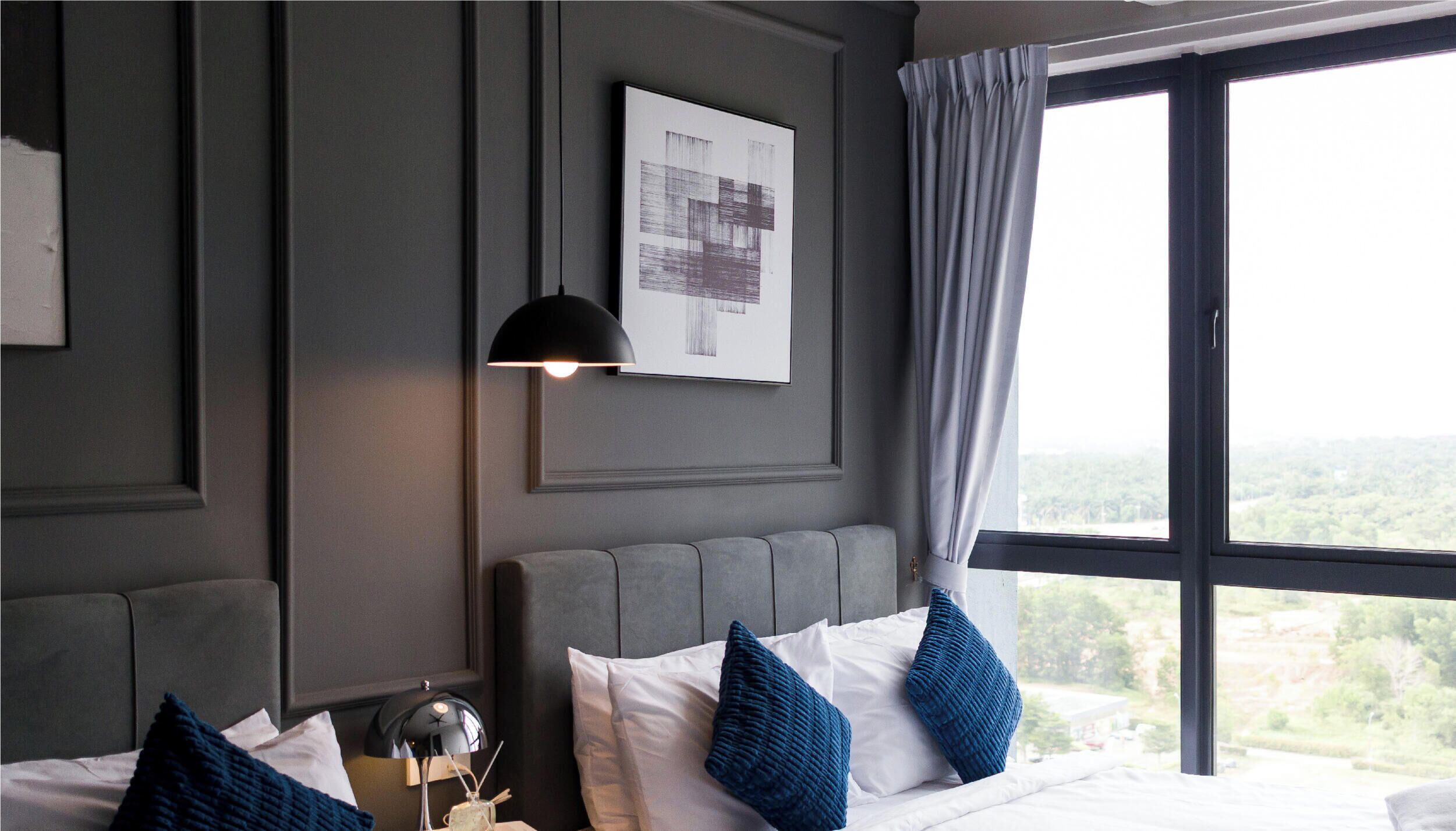 A hotel room with a large window, a bed with white and blue pillows, a wall-mounted lamp, and a framed picture.