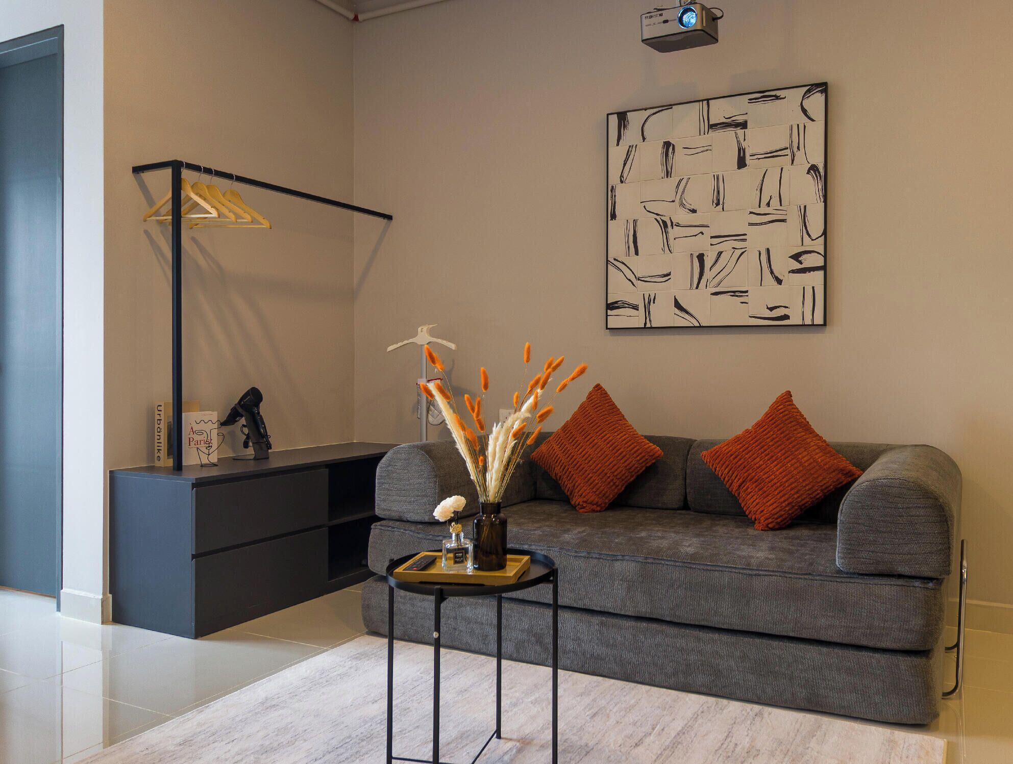 Una sala moderna con un sofá gris, dos cojines naranjas, una mesita auxiliar con un jarrón y una botella, una lámpara de pie negra y una obra de arte abstracto colgada en la pared.
