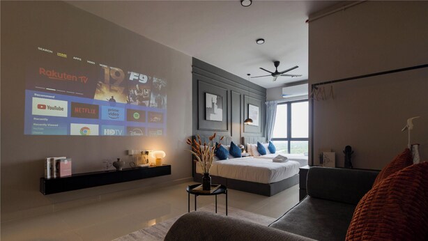 Una habitación de hotel moderna con una pantalla grande que muestra la interfaz de un servicio de streaming, una cama con vistas, un ventilador de techo y una decoración minimalista.