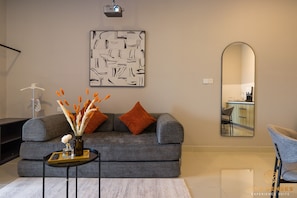 A modern living room with a grey sofa, a coffee table, a vase with decorative elements, a large abstract painting, and a mirror reflecting a kitchen area.
