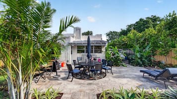 A patio with a round table and chairs, surrounded by plants and a house in the background.