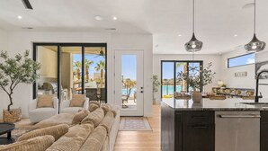 Et moderne køkken med en central ø, et spiseområde med udsigt til poolen, og en stue med en stor sofa og en plante.