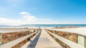 Une promenade en bois qui mène à une plage au rivage sablonneux, sous un ciel bleu et dégagé.