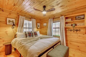 A wooden cabin bedroom with a bed, a ceiling fan, a window with curtains, and a shelf with decorative items.