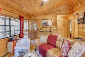 A wooden cabin interior with a leather sofa, a white armchair, a small table with a lamp, and a painting on the wall.