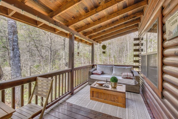 A log cabin with a wooden deck, a sofa with cushions, a coffee table, and a view of the forest.