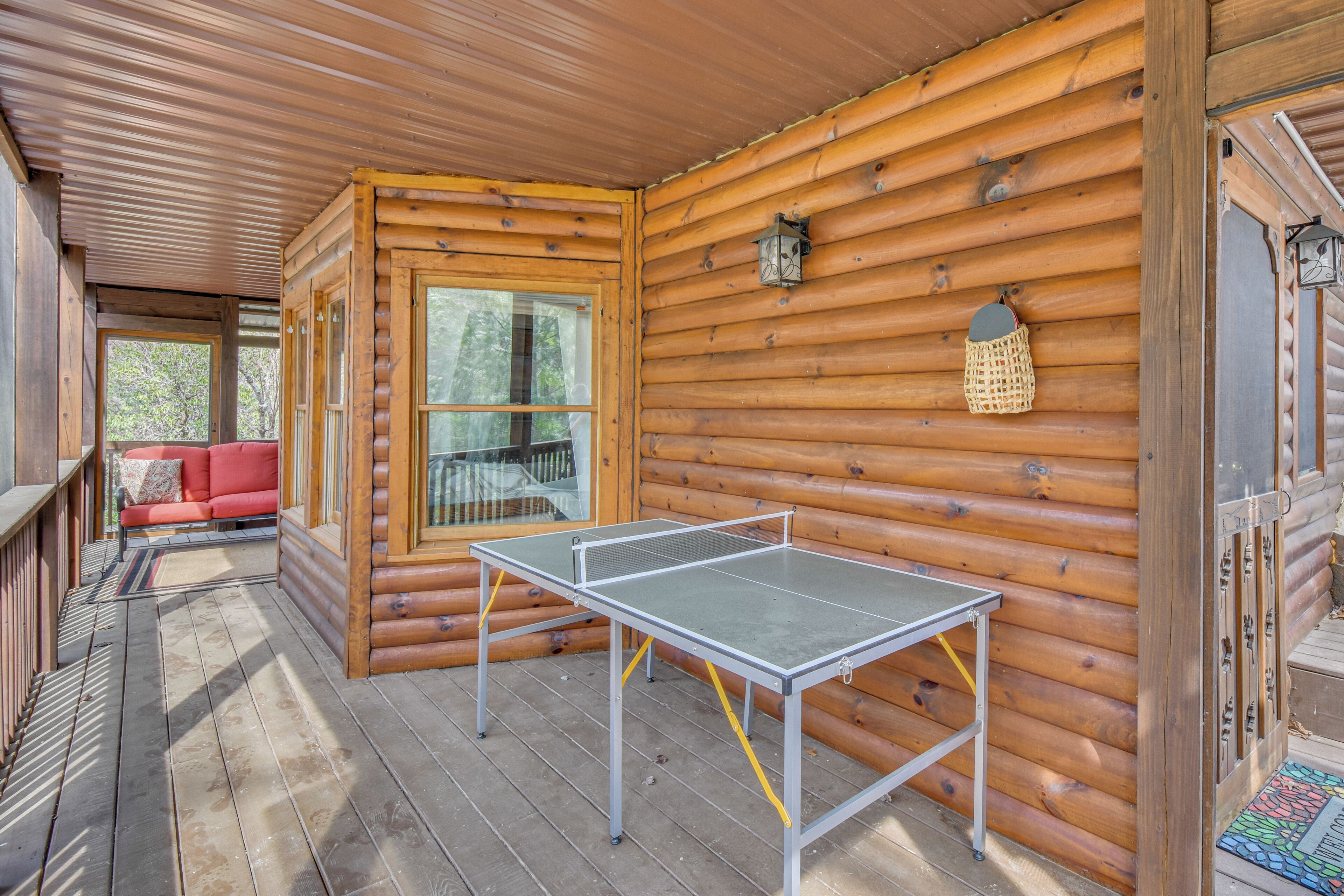 A wooden cabin with a ping pong table, a red sofa, and a window overlooking greenery.