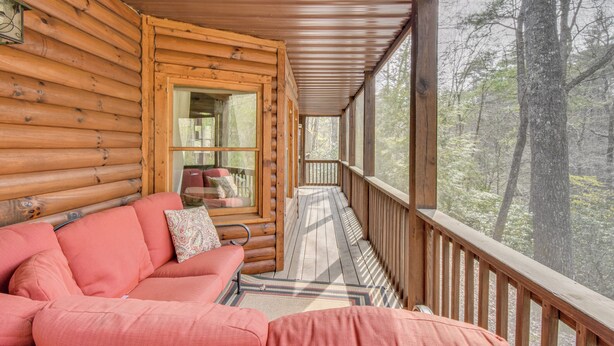 A log cabin with a wooden deck, a sofa with cushions, and a view of the forest.