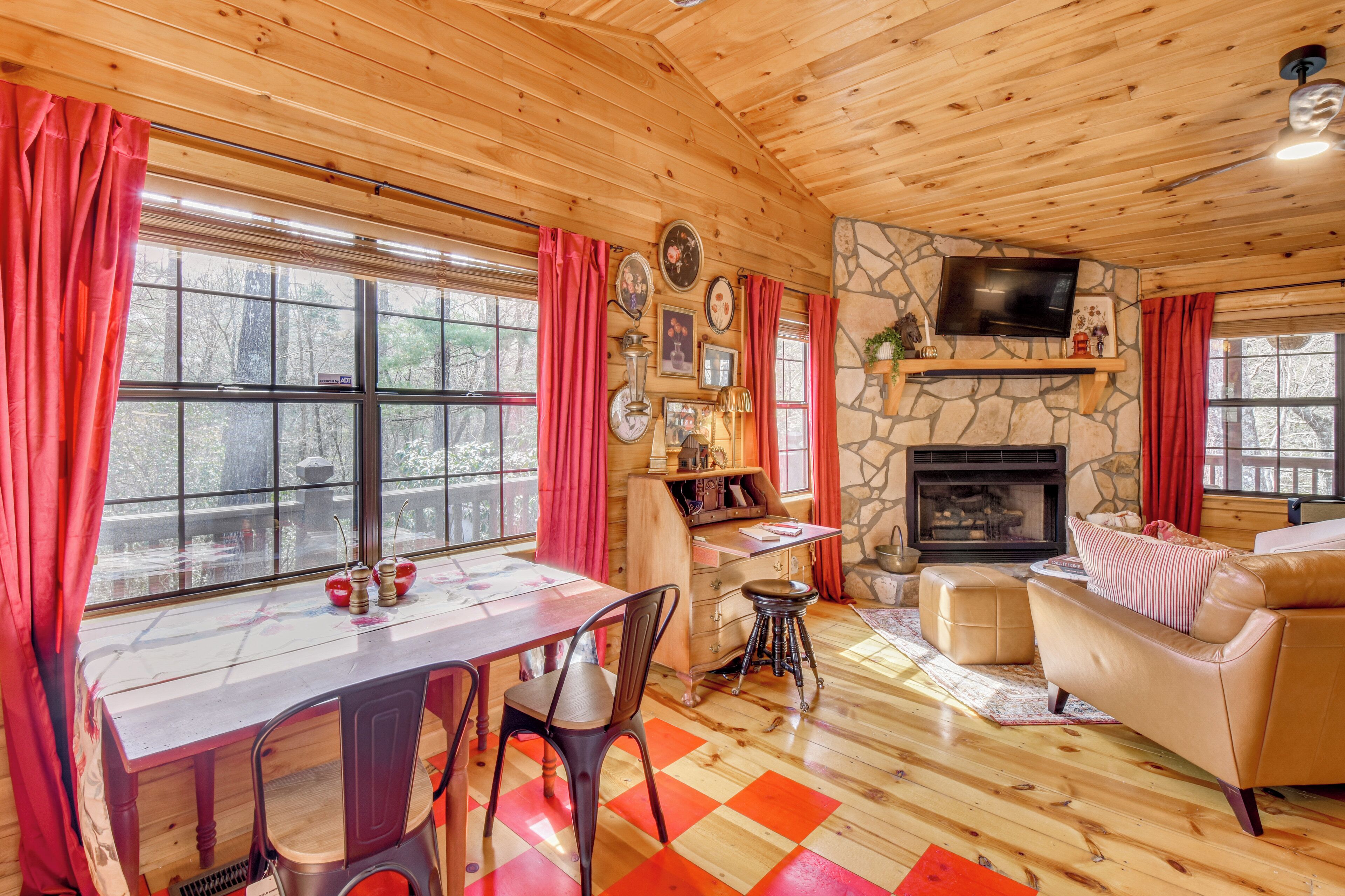 A cozy cabin interior with a wooden ceiling, stone fireplace, dining table, and leather sofa.