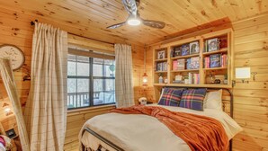 A wooden cabin bedroom with a bed, a ceiling fan, a window with curtains, and a bookshelf.