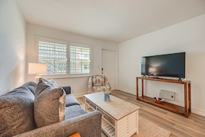 Ein Wohnzimmer mit Sofa, Sessel, Couchtisch, TV-Schrank und einem Fenster mit Jalousien.