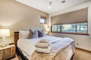 Ein Schlafzimmer mit Bett, Kissen, einem Nachttisch mit Lampe, einem Fenster mit Jalousien und Blick auf Bäume.