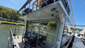 Un barco con cubierta superior acristalada que incluye una mesa, sillas, barandilla y vistas al agua y a barcos amarrados.