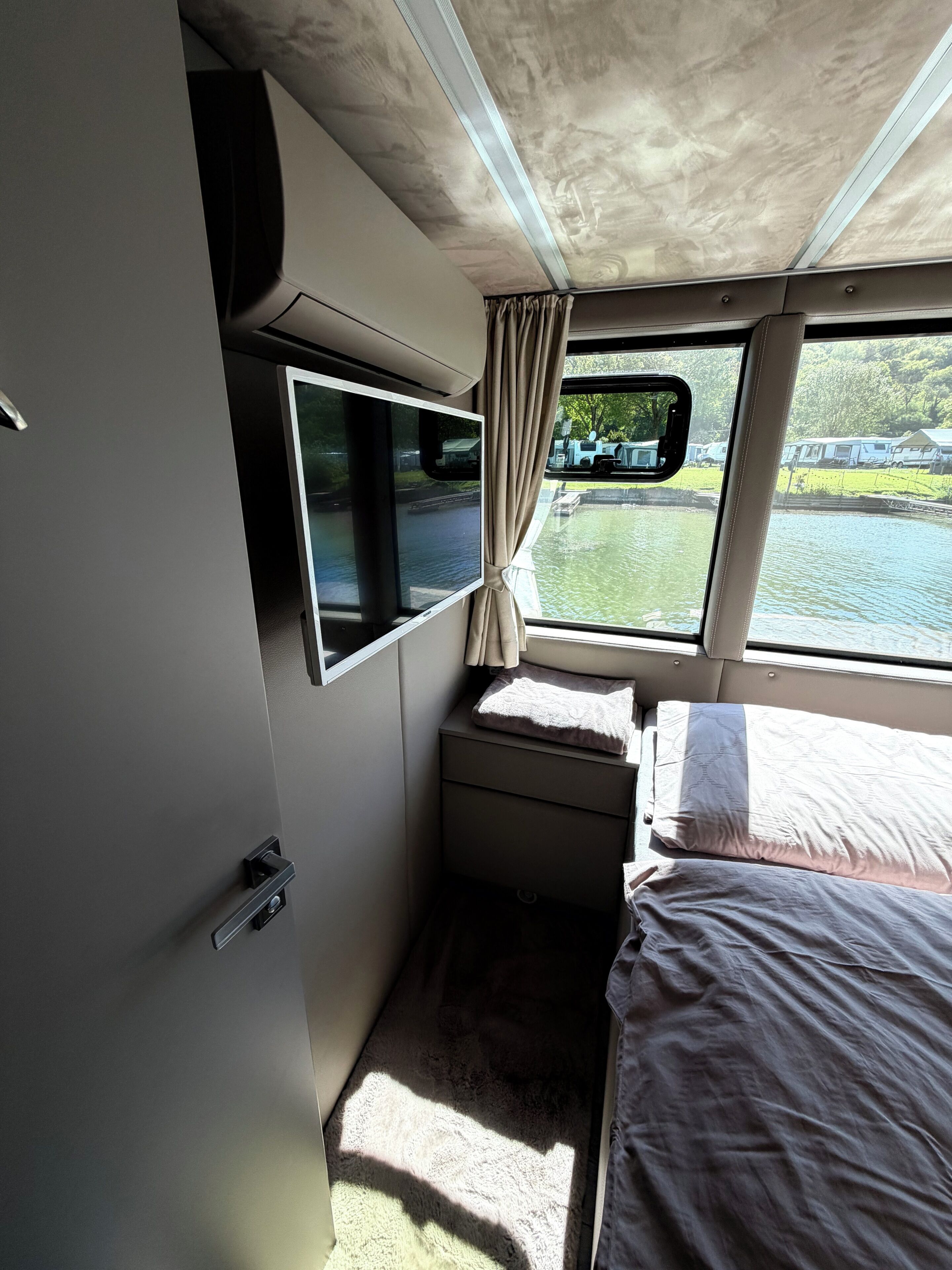 Uma cabana compacta com cama, TV e janela com vista para um lago e trailers.