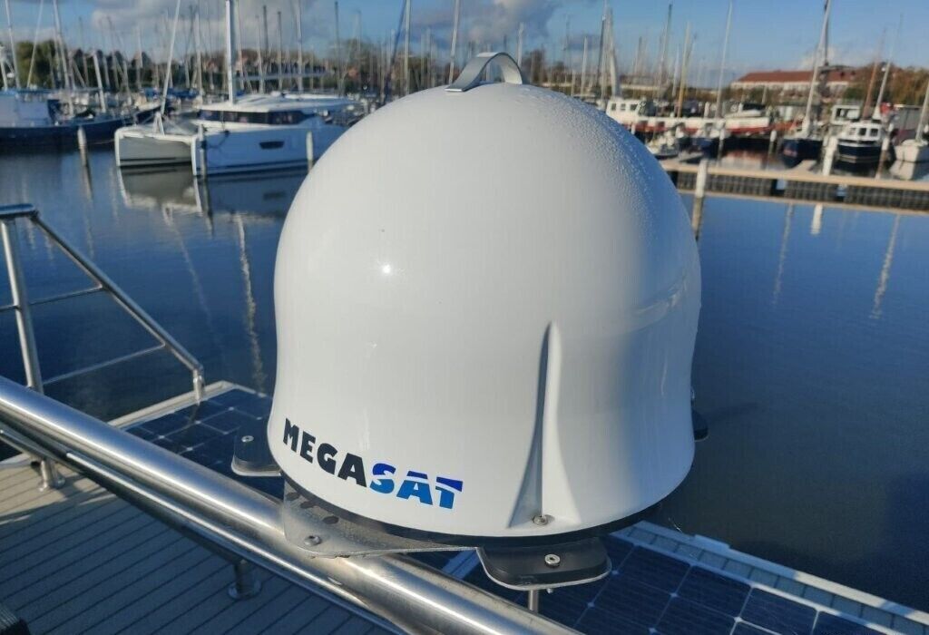 Un objeto en forma de cúpula blanca con la etiqueta 'MEGASAT' sobre la cubierta de un barco, con paneles solares y un puerto deportivo al fondo.