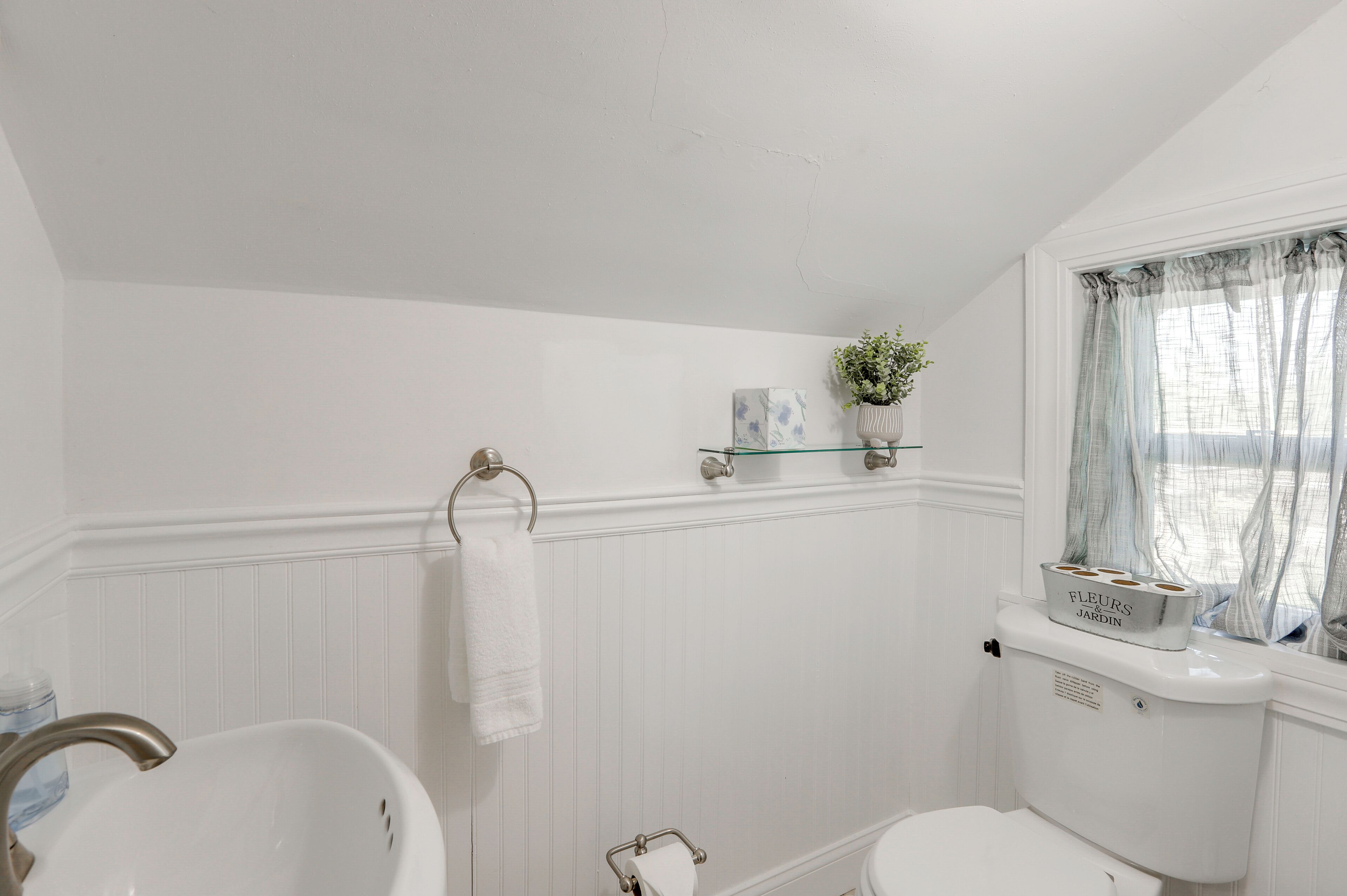 A bathroom with a white sink, toilet, and bathtub. There is a window with sheer curtains and a small potted plant on the windowsill.