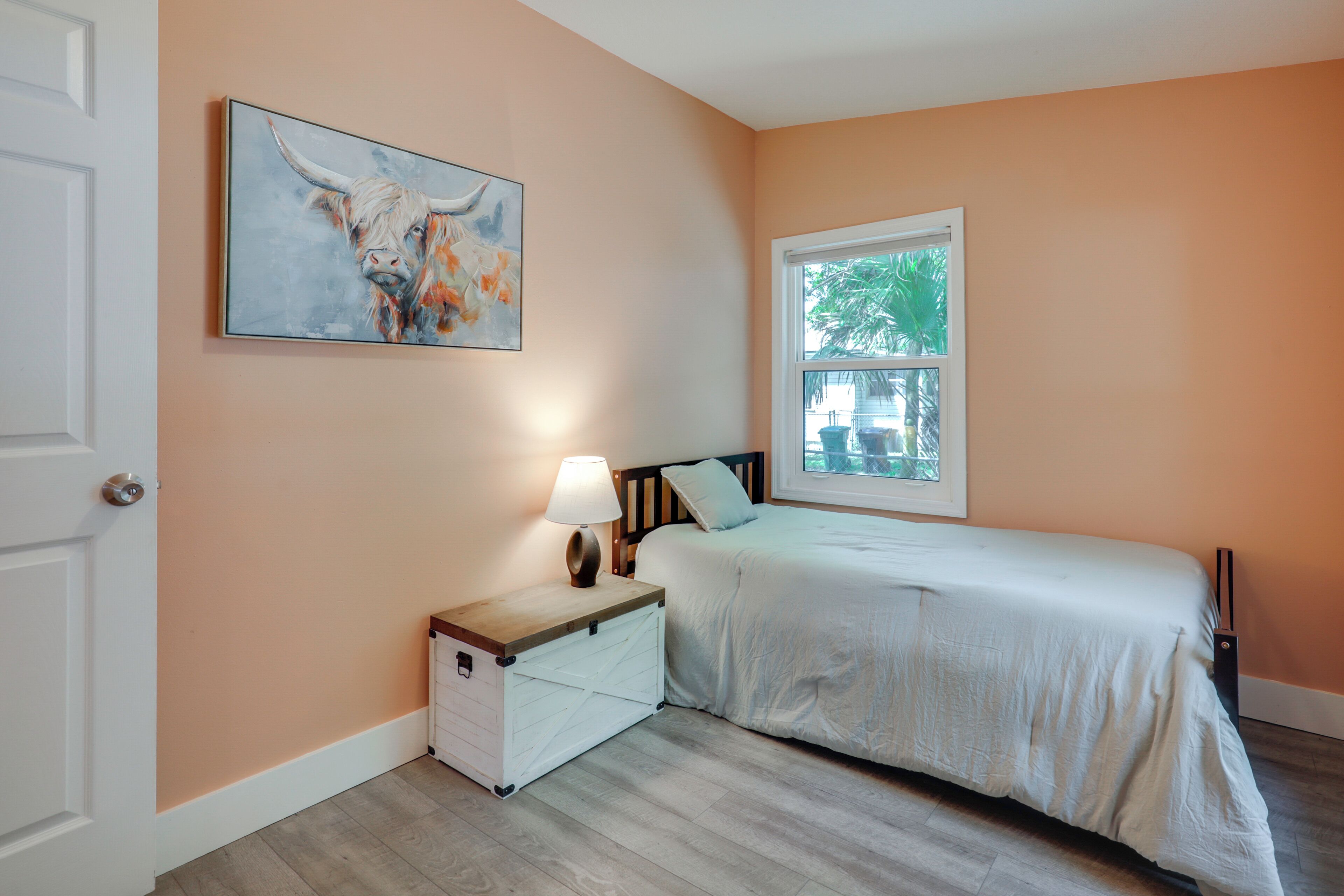 A bedroom with a bed, a nightstand with a lamp, a dresser, a window with a view, and a painting on the wall.